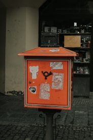A vintage orange mailbox stands on a cobblestone street, adorned with various stickers and weathered paper remnants. The backdrop features the entrance of a shop with a partially visible display and a yellow sign indicating a 30% discount.