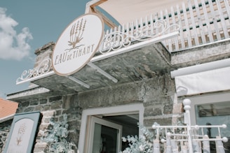 A rustic stone building with a white and elegant sign reading 'EAU de Tihany' decorated with lavender imagery. The building features a wooden balcony with a white railing above, and floral arrangements are seen below the sign.