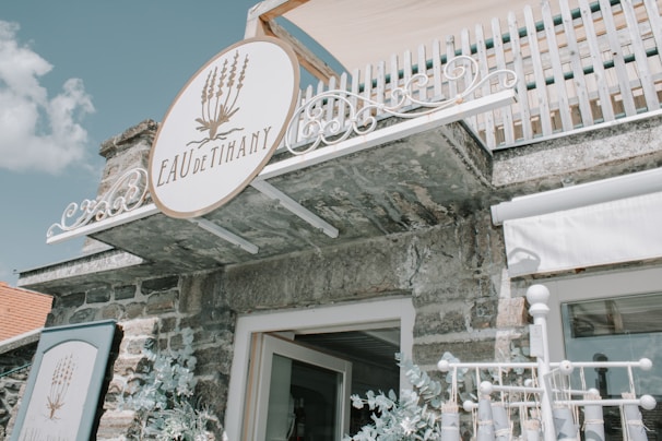 A rustic stone building with a white and elegant sign reading 'EAU de Tihany' decorated with lavender imagery. The building features a wooden balcony with a white railing above, and floral arrangements are seen below the sign.