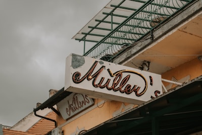 A vintage-style sign with the word 'Mullen's' in a cursive font is mounted on the side of a worn building. The sign appears slightly weathered, with some paint chipping off. Above the sign is a railing or balcony structure, with a greenish tint. The overall scene looks aged, under a cloudy sky.