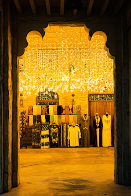 An arched entrance frames a richly decorated interior with hanging lights casting a warm yellow glow. Mannequins dressed in traditional attire stand next to a display of colorful textiles hanging on the wall.