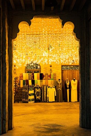 An arched entrance frames a richly decorated interior with hanging lights casting a warm yellow glow. Mannequins dressed in traditional attire stand next to a display of colorful textiles hanging on the wall.