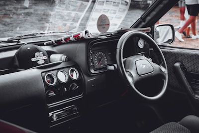 A vintage car's interior featuring a classic design with a focus on the steering wheel, dashboard, and center console. The dashboard includes round dials, and the center console has several control knobs. A black cap with the logo 'RS Owners Club' is placed on the dashboard alongside a rolled-up magazine.