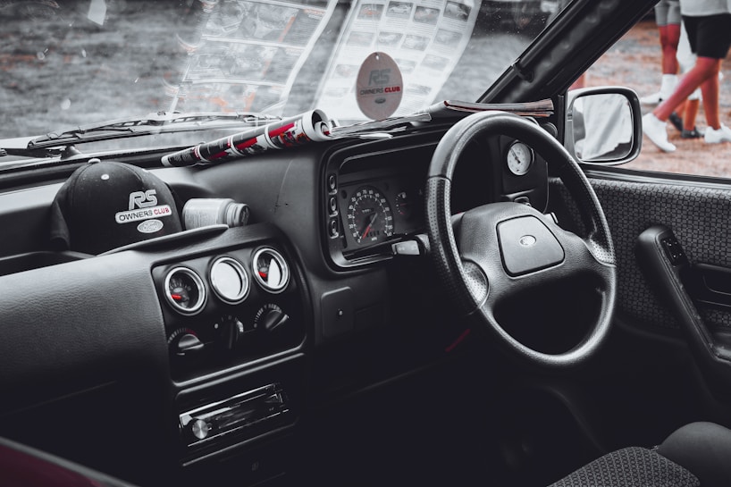 A vintage car's interior featuring a classic design with a focus on the steering wheel, dashboard, and center console. The dashboard includes round dials, and the center console has several control knobs. A black cap with the logo 'RS Owners Club' is placed on the dashboard alongside a rolled-up magazine.