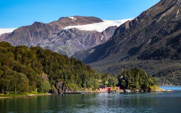 A serene fjord landscape with towering mountains in the background, partially covered by a white glacier. A small cluster of red houses sits on the edge of the water, surrounded by dense green forest. The calm water reflects the greenery and the blue sky above.
