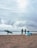 On a sandy beach, several people are engaged in activities related to surfing. One person walks with a green surfboard under their arm, while two others stand near a blue and white sail, likely for windsurfing, chatting with each other. Another individual is seen in the background holding a surfboard near the water's edge. The sky is cloudy, creating a moody atmosphere.