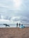 On a sandy beach, several people are engaged in activities related to surfing. One person walks with a green surfboard under their arm, while two others stand near a blue and white sail, likely for windsurfing, chatting with each other. Another individual is seen in the background holding a surfboard near the water's edge. The sky is cloudy, creating a moody atmosphere.