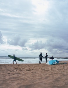 On a sandy beach, several people are engaged in activities related to surfing. One person walks with a green surfboard under their arm, while two others stand near a blue and white sail, likely for windsurfing, chatting with each other. Another individual is seen in the background holding a surfboard near the water's edge. The sky is cloudy, creating a moody atmosphere.