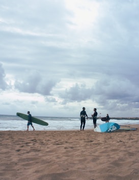 On a sandy beach, several people are engaged in activities related to surfing. One person walks with a green surfboard under their arm, while two others stand near a blue and white sail, likely for windsurfing, chatting with each other. Another individual is seen in the background holding a surfboard near the water's edge. The sky is cloudy, creating a moody atmosphere.