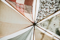 Various fabric panels with intricate patterns and earthy tones are stretched out and attached to wooden poles, creating a visually appealing canopy. The fabrics feature geometric and floral designs, adding an artistic flair.