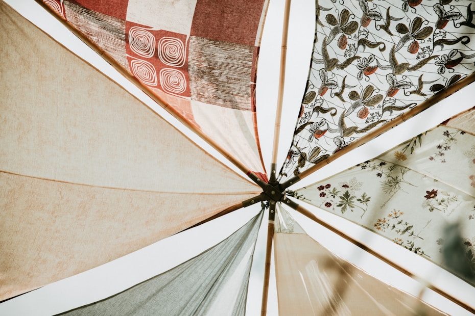 Various fabric panels with intricate patterns and earthy tones are stretched out and attached to wooden poles, creating a visually appealing canopy. The fabrics feature geometric and floral designs, adding an artistic flair.