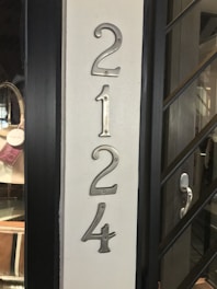 Four metallic numbers are mounted vertically on a light-colored wall. The numbers 2, 1, 2, and 4 are attached with visible screws. To the left, part of a glass door display with bags is visible, while to the right is a black frame with a modern handle.