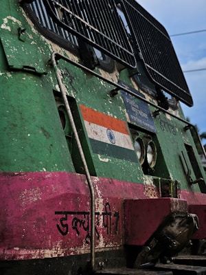 A close-up view of the front section of a weathered train engine, featuring the Indian national flag and text in Hindi. The surface is painted in green and pink, showing signs of rust and wear. Two headlights and a metal grille are visible, with some industrial fittings and components.