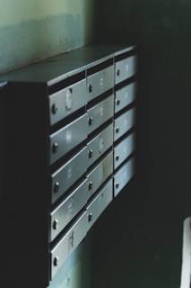 A series of metal mailboxes are mounted on a wall, arranged in two vertical columns. The mailboxes have a slightly aged and worn appearance, with some visible scratches and rust. The lighting is dim, casting shadows and giving a sense of a quiet, possibly neglected space.