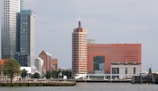 A waterfront cityscape featuring a row of modern high-rise buildings. The tallest structure is a sleek glass skyscraper with vertical lines, accompanied by a distinctive cylindrical building with horizontal red and beige stripes. In the middle ground, a large rectangular building with an orange facade and numerous small windows is present. Trees and greenery line the waterfront, with some small boats docked along the shore.