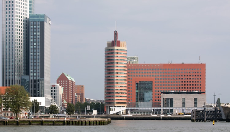 A waterfront cityscape featuring a row of modern high-rise buildings. The tallest structure is a sleek glass skyscraper with vertical lines, accompanied by a distinctive cylindrical building with horizontal red and beige stripes. In the middle ground, a large rectangular building with an orange facade and numerous small windows is present. Trees and greenery line the waterfront, with some small boats docked along the shore.