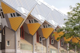 Cube-shaped housing structures with slanted roofs, displaying modern architectural design. The buildings feature a combination of yellow, gray, and brick materials, with multiple windows and geometric patterns. A tree partially obscures one of the buildings on the right side.