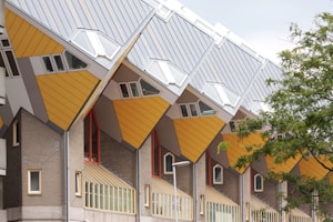Cube-shaped housing structures with slanted roofs, displaying modern architectural design. The buildings feature a combination of yellow, gray, and brick materials, with multiple windows and geometric patterns. A tree partially obscures one of the buildings on the right side.
