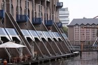 A waterfront scene with a row of modern, apartment buildings featuring distinct slanted roofs and blue accents. There is an outdoor dining area with several tables and chairs under large umbrellas near the water's edge. In the background, another building with large windows and architectural details is visible.
