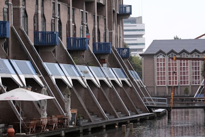 A waterfront scene with a row of modern, apartment buildings featuring distinct slanted roofs and blue accents. There is an outdoor dining area with several tables and chairs under large umbrellas near the water's edge. In the background, another building with large windows and architectural details is visible.
