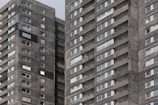 A high-rise residential building with dark gray brick facade and numerous windows. The structure features repeating patterns of balconies and windows, providing a uniform appearance. One person can be seen on a balcony, offering a sense of scale and activity.