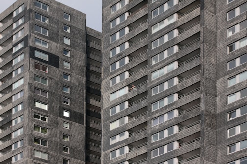 A high-rise residential building with dark gray brick facade and numerous windows. The structure features repeating patterns of balconies and windows, providing a uniform appearance. One person can be seen on a balcony, offering a sense of scale and activity.