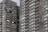 A high-rise residential building with dark gray brick facade and numerous windows. The structure features repeating patterns of balconies and windows, providing a uniform appearance. One person can be seen on a balcony, offering a sense of scale and activity.