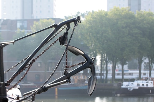 An anchor attached to a ship with chains is prominently displayed in the foreground. In the background, there are trees, buildings, and a body of water with a docked boat. The scene suggests an industrial or maritime setting.