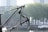 An anchor attached to a ship with chains is prominently displayed in the foreground. In the background, there are trees, buildings, and a body of water with a docked boat. The scene suggests an industrial or maritime setting.