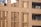 The image features a modern building facade with a brick pattern. The structure includes large windows and balconies with potted plants and small decorative objects. The architecture emphasizes vertical lines and a grid-like design using beige and brown tones.