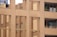 The image features a modern building facade with a brick pattern. The structure includes large windows and balconies with potted plants and small decorative objects. The architecture emphasizes vertical lines and a grid-like design using beige and brown tones.
