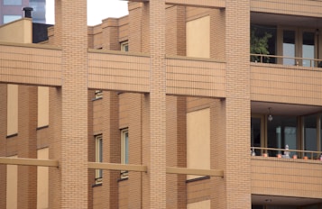 The image features a modern building facade with a brick pattern. The structure includes large windows and balconies with potted plants and small decorative objects. The architecture emphasizes vertical lines and a grid-like design using beige and brown tones.
