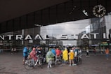 A group of people wearing colorful rain ponchos gather outside a large modern building with a glass facade. Above the entrance, large letters spell out 'CENTRAAL STATION'. A large clock is mounted on the upper right corner of the building. Several people are standing next to bicycles, and there are other pedestrians nearby.
