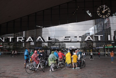 A group of people wearing colorful rain ponchos gather outside a large modern building with a glass facade. Above the entrance, large letters spell out 'CENTRAAL STATION'. A large clock is mounted on the upper right corner of the building. Several people are standing next to bicycles, and there are other pedestrians nearby.