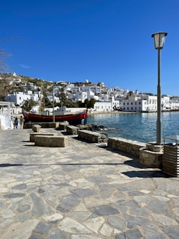 A coastal town with whitewashed buildings and a rocky shoreline. The clear blue sky enhances the vibrant colors of the sea, and a traditional wooden boat is docked near the shore. Stone benches and a streetlamp line the paved waterfront promenade.