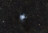A field of stars scatters across the expanse of the night sky. At the center, a bright nebula displays a turbulent mix of luminous clouds, glowing with a soft bluish-white hue. Surrounding the nebula, a dense array of stars twinkles subtly against the dark backdrop of space.