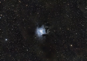 A field of stars scatters across the expanse of the night sky. At the center, a bright nebula displays a turbulent mix of luminous clouds, glowing with a soft bluish-white hue. Surrounding the nebula, a dense array of stars twinkles subtly against the dark backdrop of space.