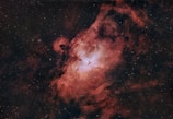 A stunning cosmic scene featuring a nebula with swirling clouds of red and pink gas and dust, illuminated by bright stars. The central region appears more luminous, with a mixture of white and pale blue hues, suggesting areas of intense energy and formation.