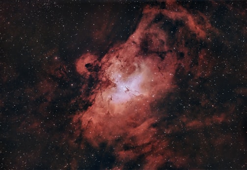 A stunning cosmic scene featuring a nebula with swirling clouds of red and pink gas and dust, illuminated by bright stars. The central region appears more luminous, with a mixture of white and pale blue hues, suggesting areas of intense energy and formation.