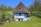 A rustic, thatched-roof cottage surrounded by lush greenery and trees, situated in a rural setting. The cottage appears weathered, with a blue and white exterior, small wooden windows, and a large, steeply sloped roof. Nearby, there are wooden structures and simple fences, with a bright landscape accentuated by a clear blue sky.