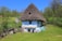 A rustic, thatched-roof cottage surrounded by lush greenery and trees, situated in a rural setting. The cottage appears weathered, with a blue and white exterior, small wooden windows, and a large, steeply sloped roof. Nearby, there are wooden structures and simple fences, with a bright landscape accentuated by a clear blue sky.