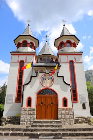 A church with two tall, pointed towers topped with crosses. The facade features red architectural accents and a religious icon above the wooden doors. Romanian flags are displayed on either side of the entrance. The structure is built on a stone foundation, and the sky is partly cloudy with patches of blue visible.