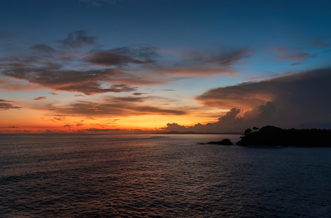 a sunset over a body of water with clouds in the sky, Breathtaking oceanfront sunset view from a tropical beach. Witness