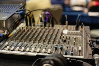 A sound mixing console with numerous faders, knobs, and indicators is prominently placed on a desk. A laptop can be seen on the left side, slightly elevated. Headphones are positioned on the surface near the mixing board, and various cables are connected to the console.