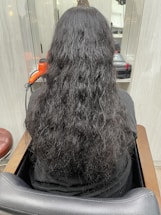 A person with long, wavy black hair is seated in a salon chair, facing away from the camera. An orange hair dryer and some salon equipment are visible on the counter in front of them.