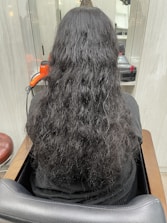 A person with long, wavy black hair is seated in a salon chair, facing away from the camera. An orange hair dryer and some salon equipment are visible on the counter in front of them.