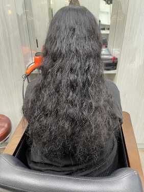 A person with long, wavy black hair is seated in a salon chair, facing away from the camera. An orange hair dryer and some salon equipment are visible on the counter in front of them.