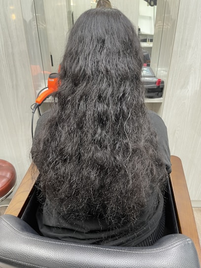A person with long, wavy black hair is seated in a salon chair, facing away from the camera. An orange hair dryer and some salon equipment are visible on the counter in front of them.
