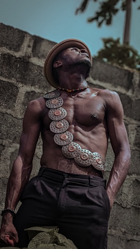A shirtless man wearing a decorative strap with metal disc embellishments across his chest stands against a textured stone wall. He is wearing a hat and looking upwards with a strong, determined expression. The lighting highlights the muscular definition of his body, creating a striking contrast with the darker tones of his pants and the wall.