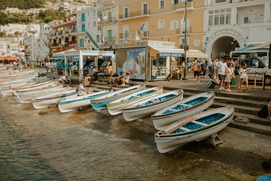 A seaside promenade features multiple small white and blue rowing boats lined up along the water's edge. People are seen walking and interacting near buildings that have a Mediterranean architectural style, painted in shades of white, yellow, and red. A sign advertising boat tours is visible next to a group of tourists, and the overall scene appears lively and bustling.